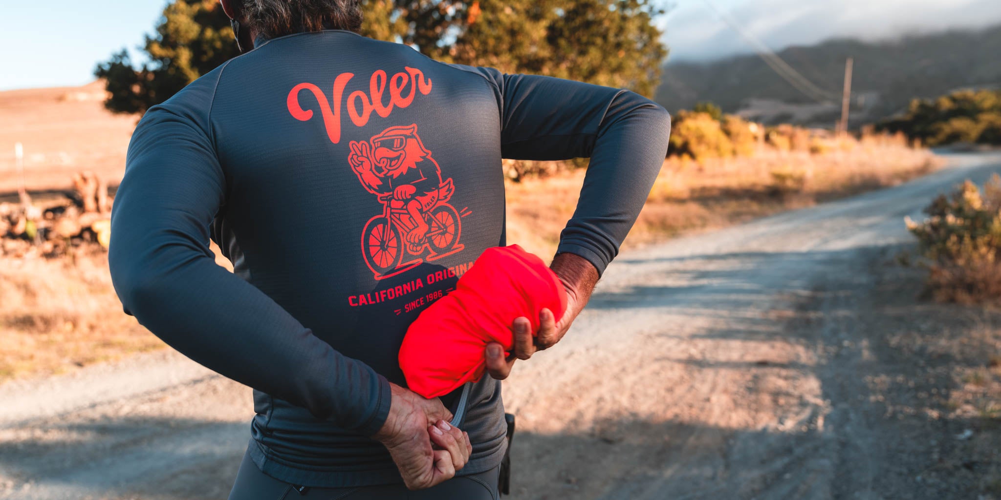 Person wearing a black long-sleeve shirt with 'Voler' branding, holding red jacket on a dirt road.