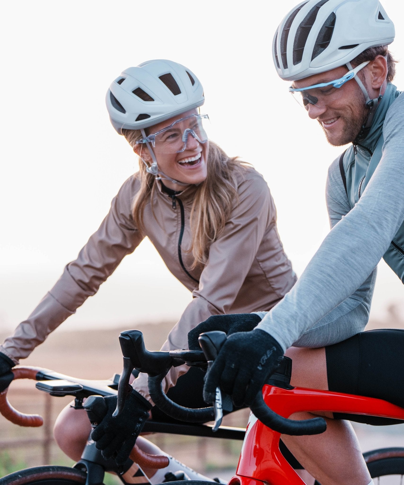 Two cyclists on a road bike with helmets and sunglasses, smiling and looking at each other.